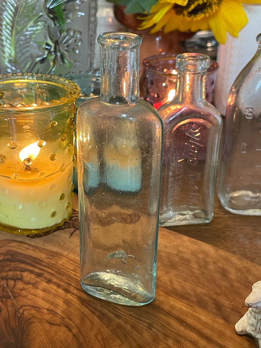 Vintage light blue oval glass pharmacy bottle shown on a lace-covered table with candlelit background. Photographed at various angles to highlight soda deposits, air bubbles, and soft molded texture.