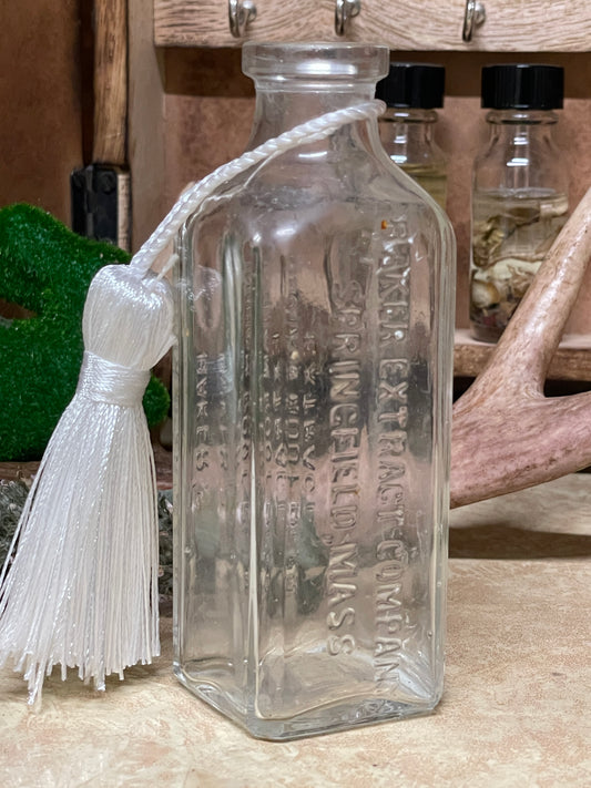Antique Baker's Indian Root Beer Extract glass bottle photographed on a lace-covered table with warm candlelight. Multiple angles show embossed lettering, square thick glass panels, and gentle neck clouding.