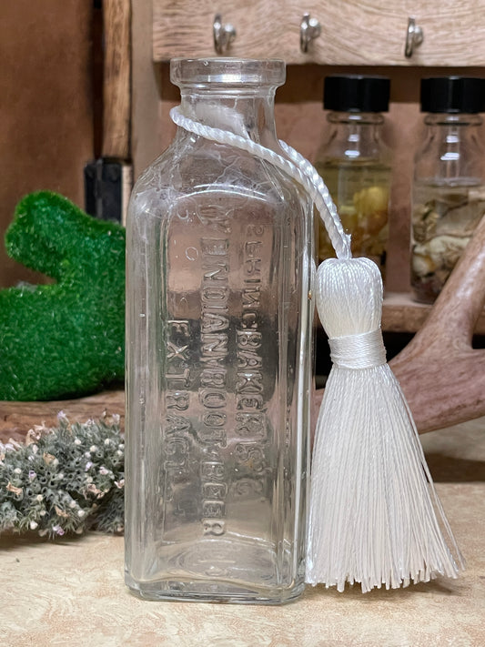 Antique Baker's Indian Root Beer Extract glass bottle photographed on a lace-covered table with warm candlelight. Multiple angles show embossed lettering, square thick glass panels, and gentle neck clouding.