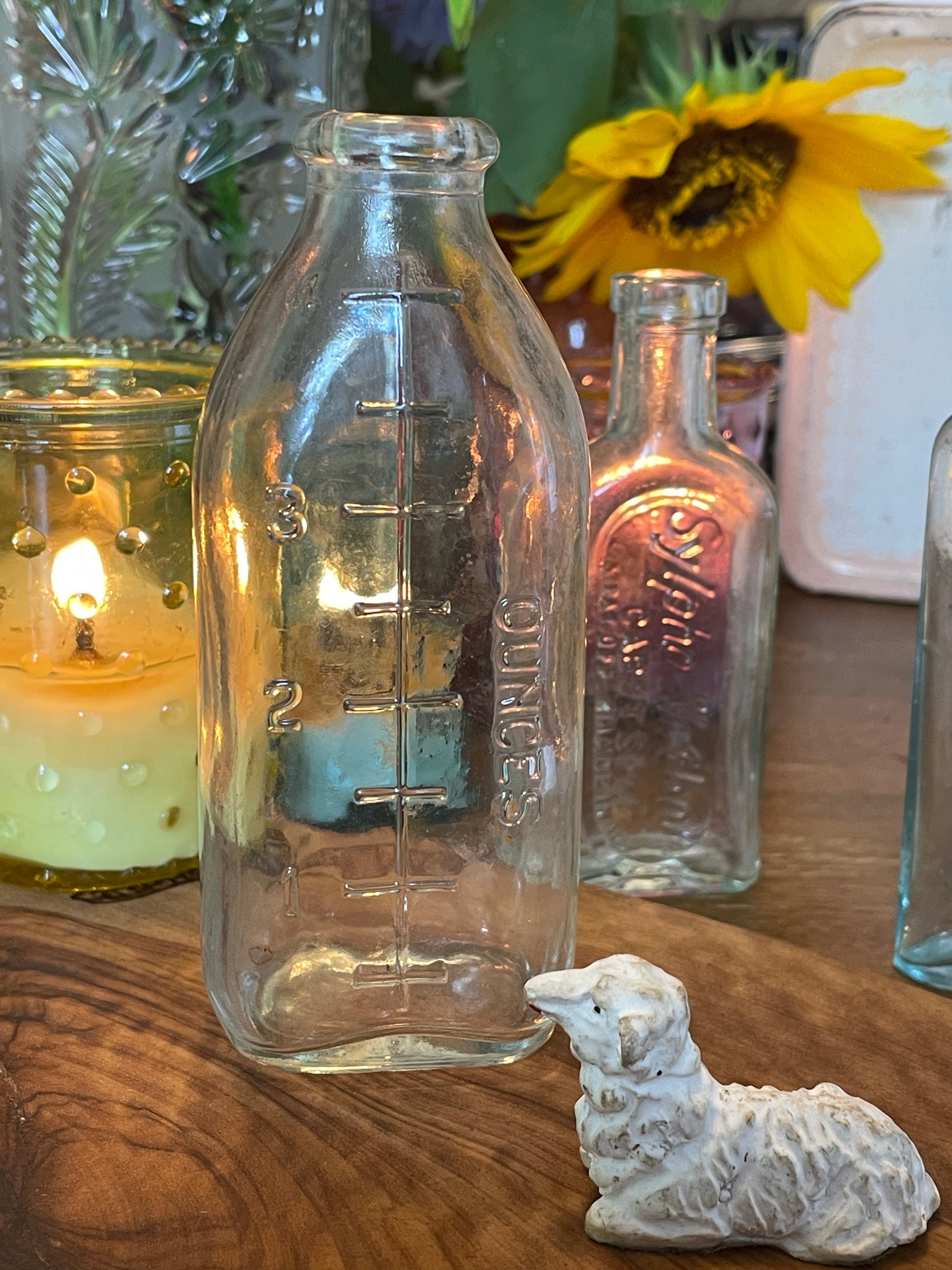Antique clear glass nursing bottle with embossed ounce measurements photographed on a lace-covered table with candlelight. Shown from multiple angles to highlight vintage markings and clarity.