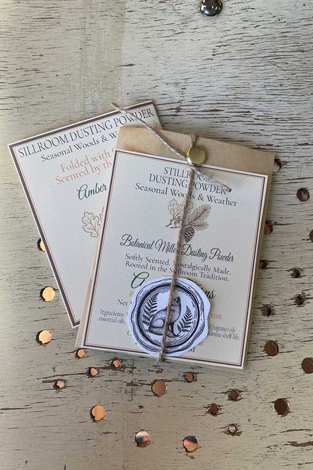 Sample Sized Dusting Powder part of the Seasonal Woods & Weather Collection. Amber Ferns Sample powder resting on a rustic wooden background with the Collection Information card peeking out.