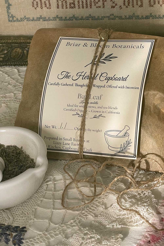 A 1 oz. package of herbs wrapped in kraft paper, tied with twine and secured with a brass fastener, sits on a vintage table draped in lace. The moody, sunlit scene includes a glimpse of a white mortar and pestle filled with dried herbs, a stray piece of twine, a sprig of lavender, and a vintage cross-stitch picture in the background, evoking a quiet stillroom atmosphere.