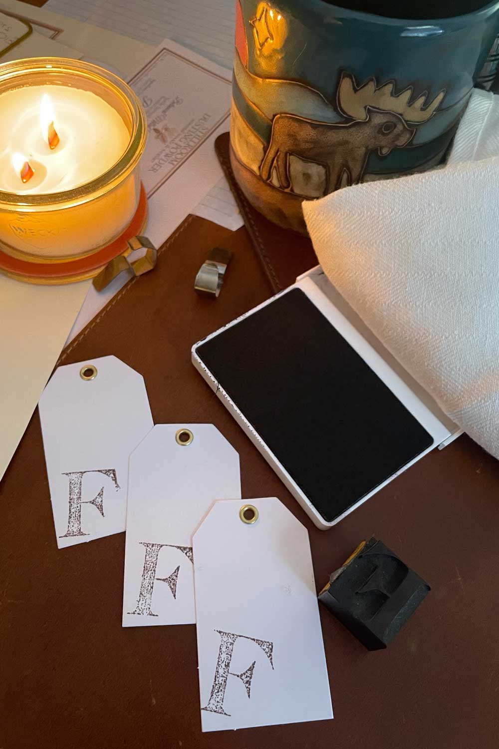 Handmade winter gift tags stamped with vintage letterpress “F” on a cozy stillroom workbench beside a beeswax candle and ink pad — part of the Midwinter Archive cottagecore collection.