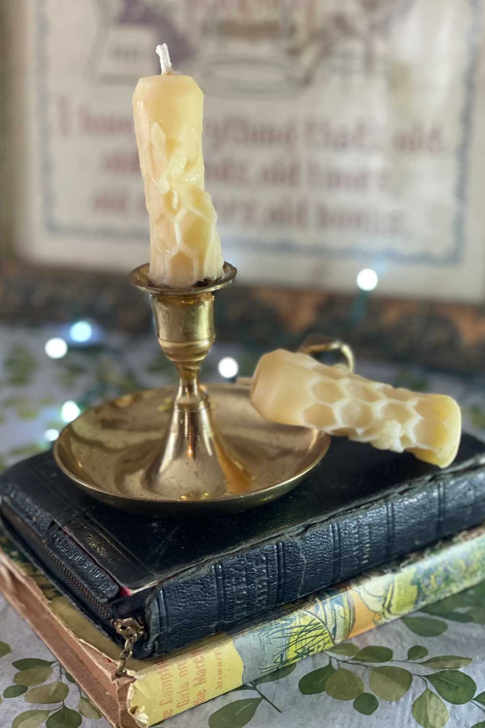 a mini beeswax bee and honeycomb taper candle sitting in a vintage brass holder on top of old books while soft lights twinkle in the background.