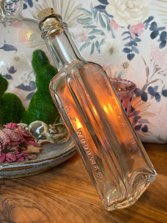 Antique Victorian-era O.F. Woodward clear glass medicine bottle shown on a lace-covered table with candlelight. Photographed at angles to reveal embossed lettering and soft clouding.