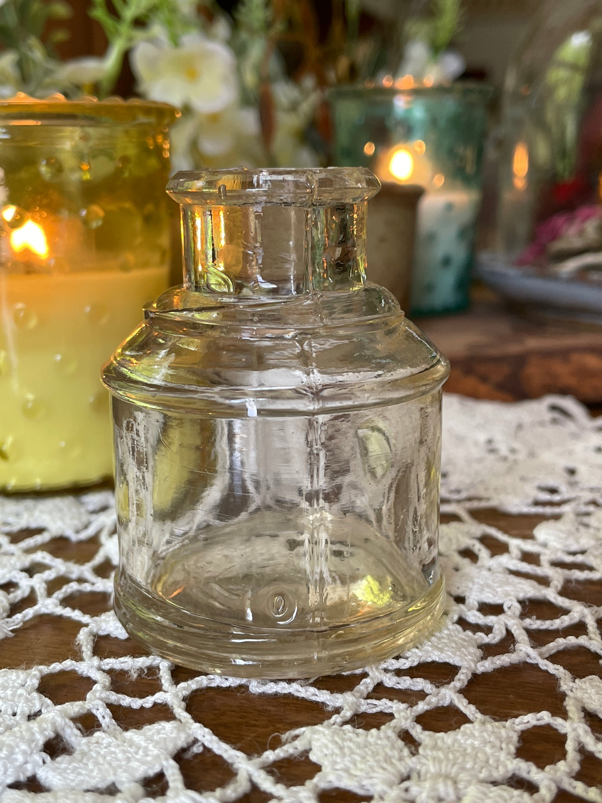 Victorian clear glass ink bottle with ribbed pen rest photographed on a vintage lace-covered table with soft candlelight. Shown at different angles to highlight bubbles in the glass and small chip detail.