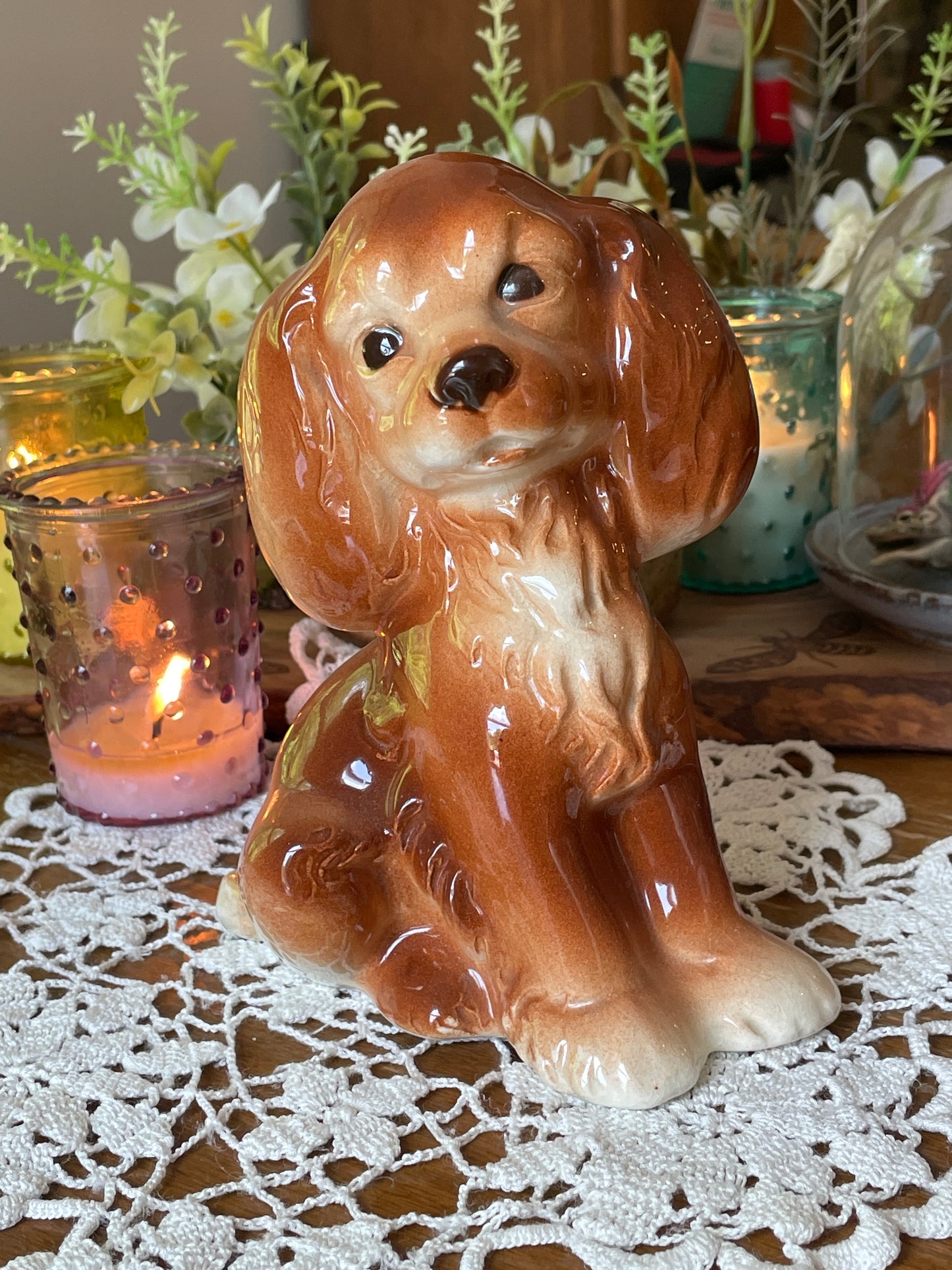 Vintage porcelain spaniel dog figurine sitting upright, photographed on a lace-covered table in soft natural light. Multiple angles show glossy finish, ears, and sweet facial expression.