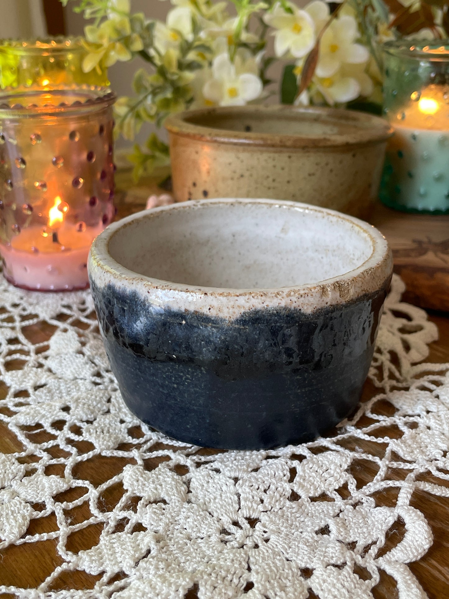 Small handmade pottery bowl with blue and white glaze shown on a lace-covered table in natural light. Photographed at various angles to highlight glaze texture and size.