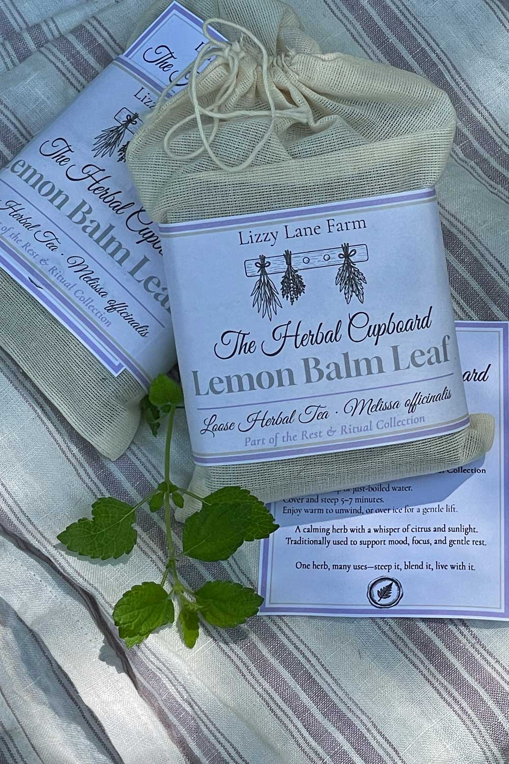 Lemon balm herbal tea in a muslin pouch with Lizzy Lane Farm label, resting on striped farmhouse linen alongside fresh lemon balm sprig.