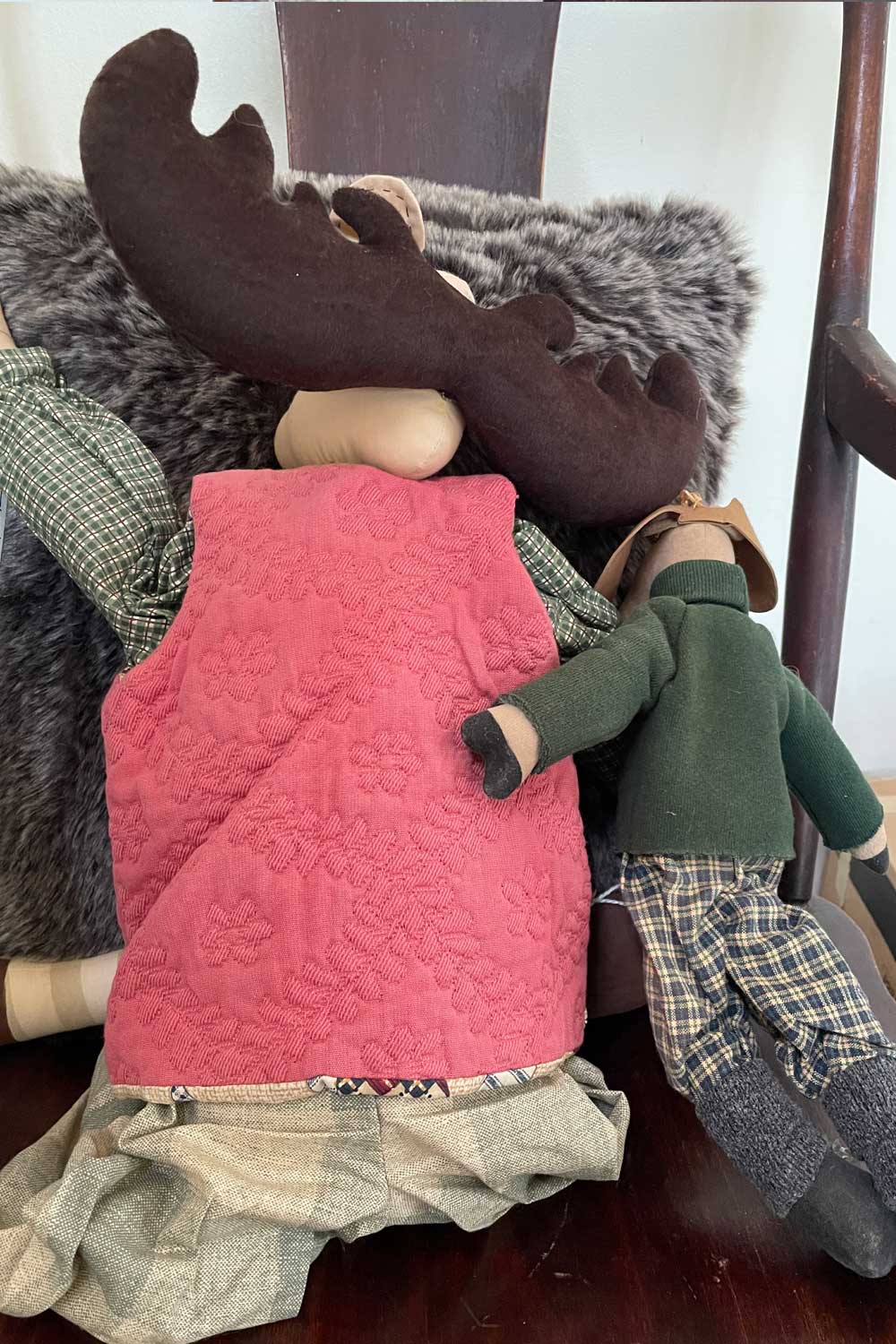 Two vintage folk art dolls, one wearing a pink quilted vest and plaid pants, the other in a green outfit with plaid pants, against a wooden chair.