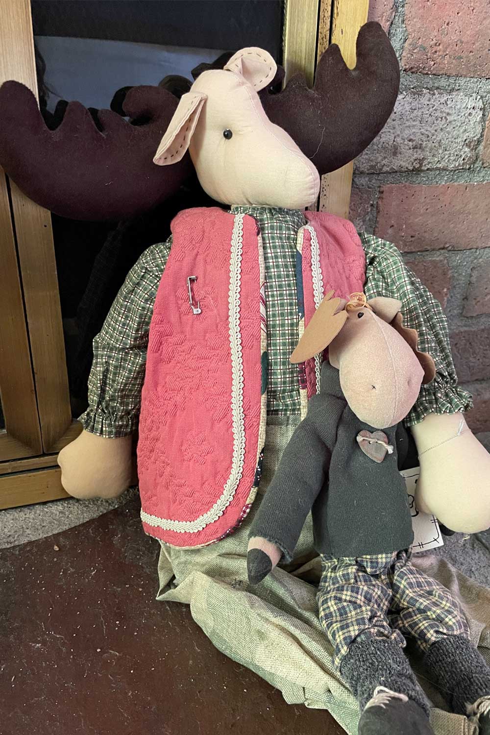 Two handmade plush moose folk art vintage dolls, one larger with a pink vest and plaid shirt, the other smaller, sitting on a wooden floor.