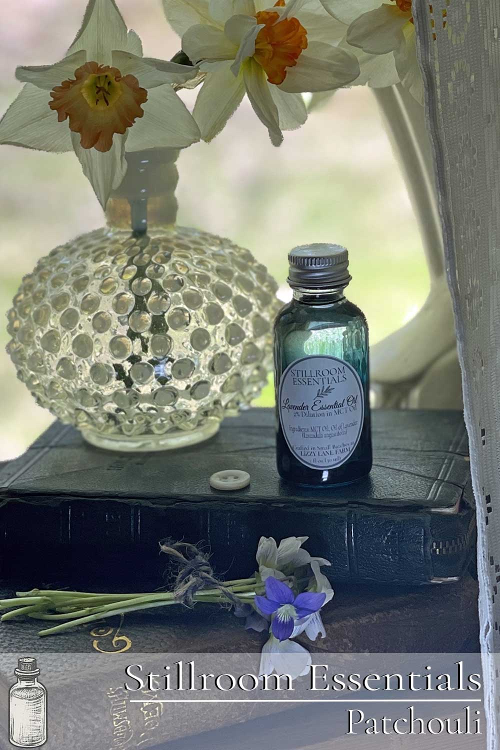 A bottle of patchouli essential oil rests on vintage books beside daffodils in a hobnail vase. A lace curtain softly veils the scene, capturing a quiet, nostalgic farmhouse moment from Lizzy Lane Farm’s Stillroom Essentials collection.