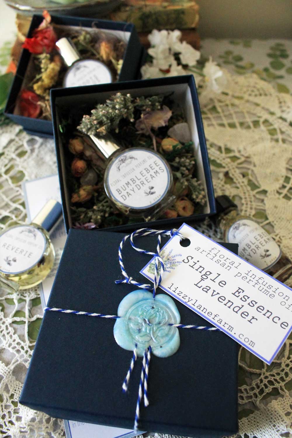 Floral Infusion Artisan Perfume Oil packaging – a deep blue gift box tied with blue baker’s twine and finished with a light blue wax seal featuring a flower. A handwritten scent tag is attached with twine. Displayed on a vintage lace doily with two open boxes in the background, revealing dried herbs and artisan perfume bottles nestled inside. Elegant, botanical, and vintage-inspired presentation.
