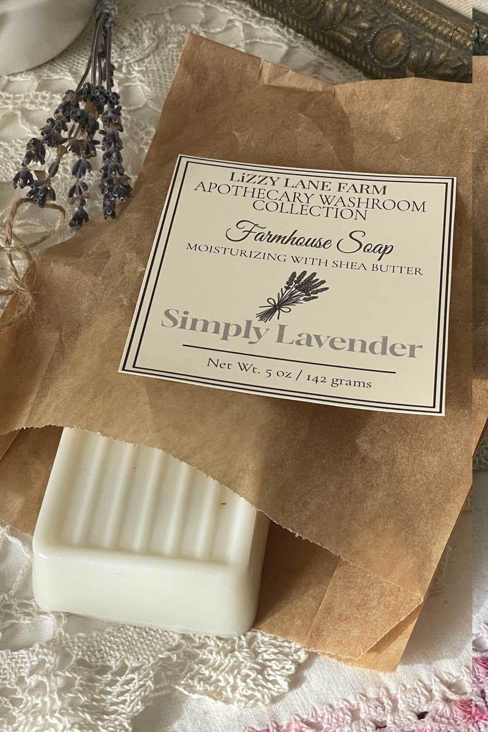 A creamy white lavender soap bar rests partially unwrapped in kraft paper, revealing its grooved surface. A curl of twine and a simple black-and-white label lie nearby. Vintage lace and ivory buttons frame the scene, with soft natural light adding warmth and a nostalgic farmhouse feel. A fully wrapped bar appears in the background, showing the full packaging.