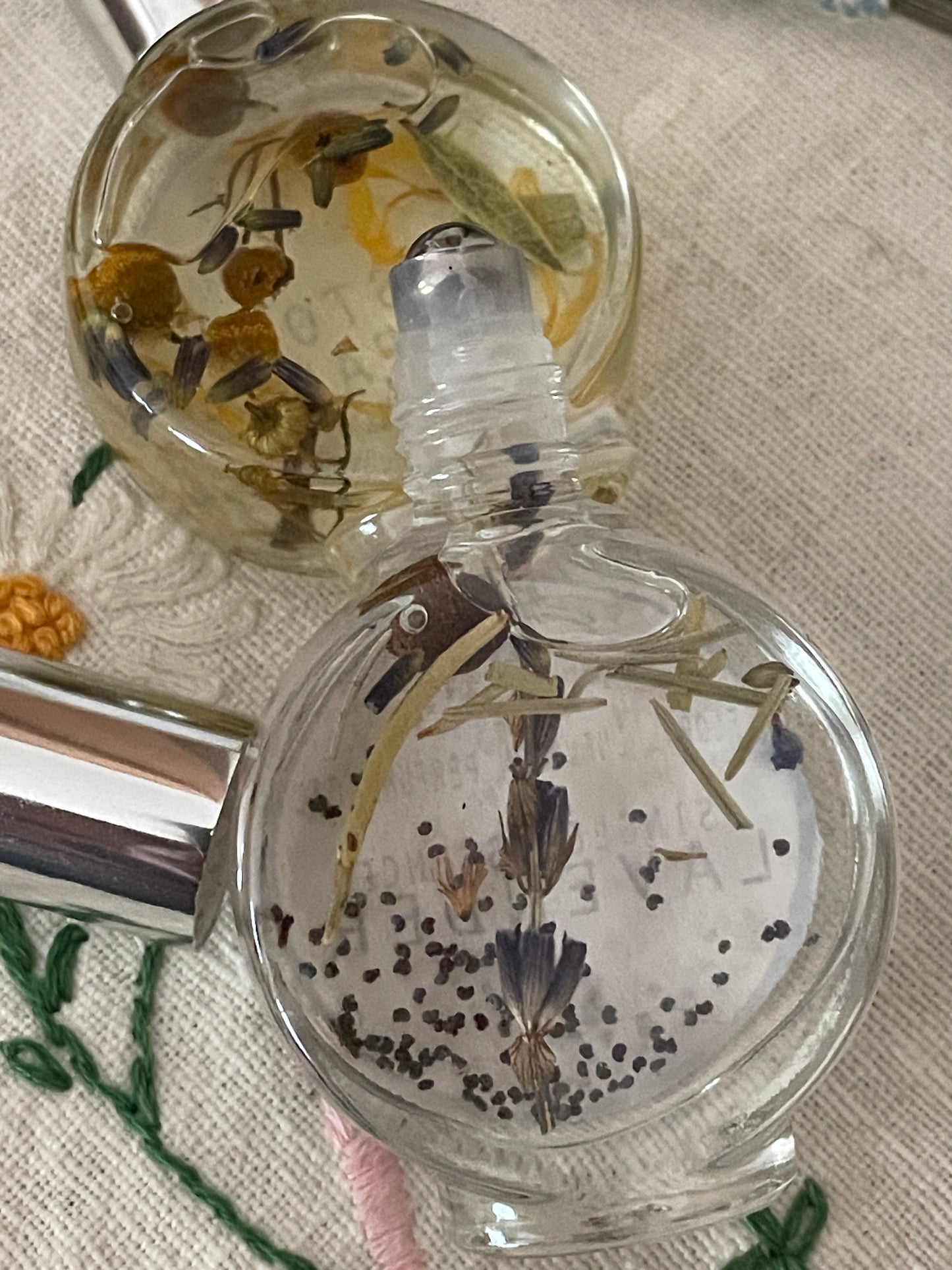 A glass perfume bottle infused with lavender spikes, seeds, and garden greenery sits atop a delicate, hand-embroidered table scarf. Beside it, a bundle of dried lavender spikes rests, their soft purple hues complementing the scene. A vintage travel clock with an aged brass finish completes the composition, evoking a sense of nostalgia and timeless elegance.