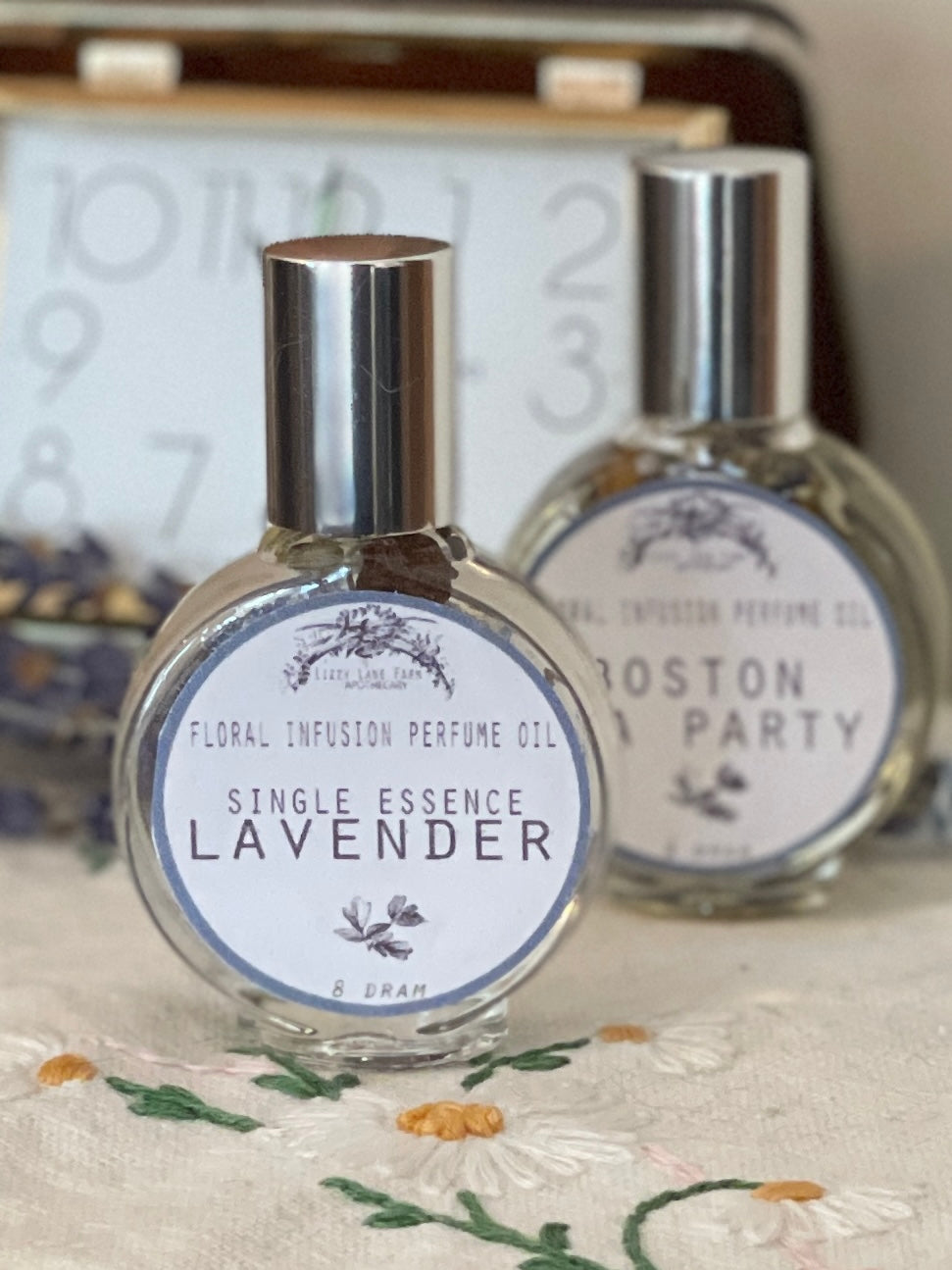 A glass perfume bottle infused with lavender spikes, seeds, and garden greenery sits atop a delicate, hand-embroidered table scarf. Beside it, a bundle of dried lavender spikes rests, their soft purple hues complementing the scene. A vintage travel clock with an aged brass finish completes the composition, evoking a sense of nostalgia and timeless elegance.