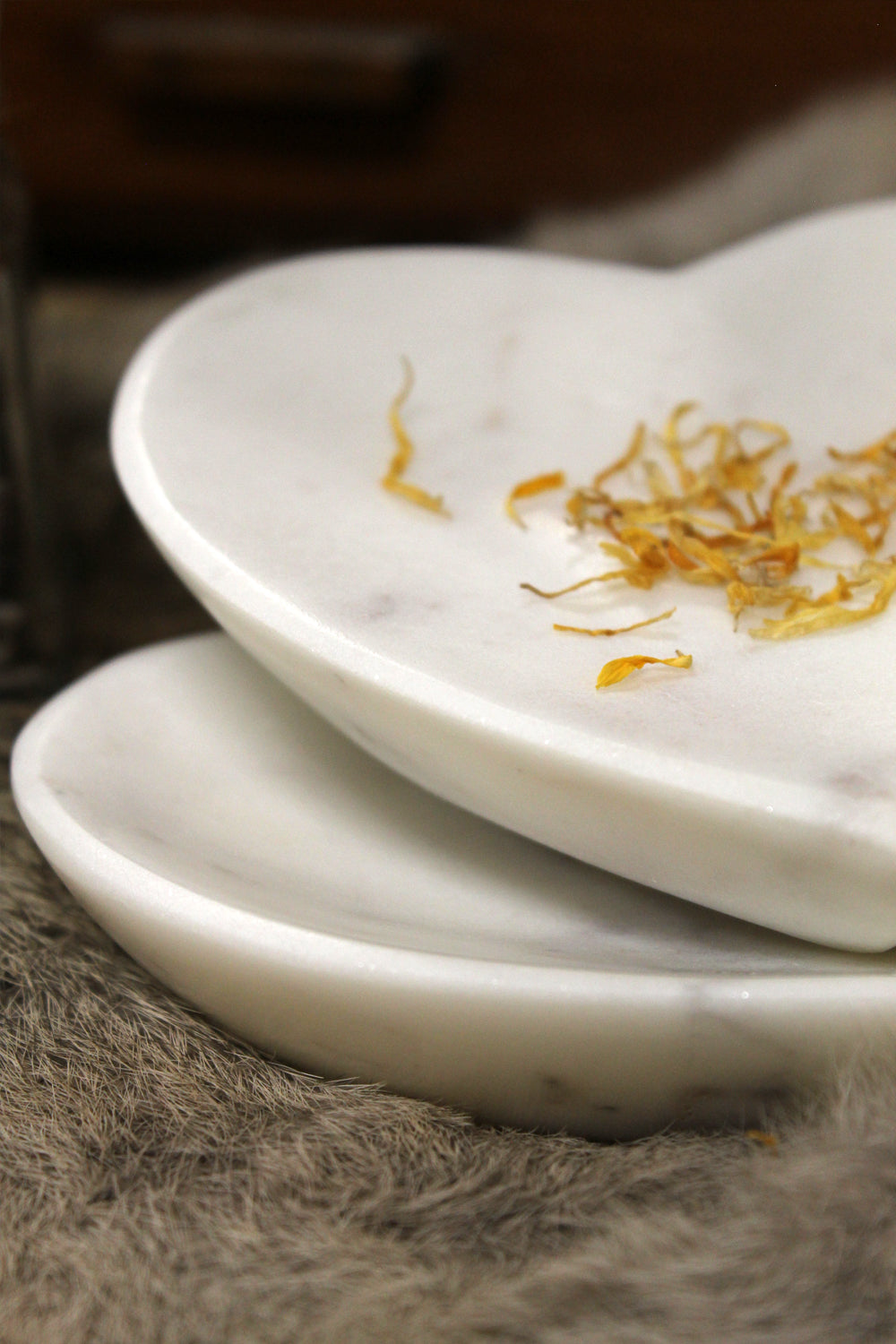 White marble heart-shaped bowl resting on soft fur, surrounded by dried petals and herbs. Natural stone veining adds warmth and organic beauty in soft light.