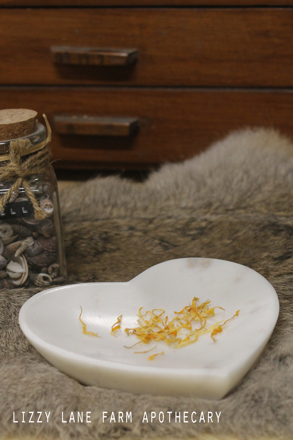 White marble heart-shaped bowl resting on soft fur, surrounded by dried petals and herbs. Natural stone veining adds warmth and organic beauty in soft light.