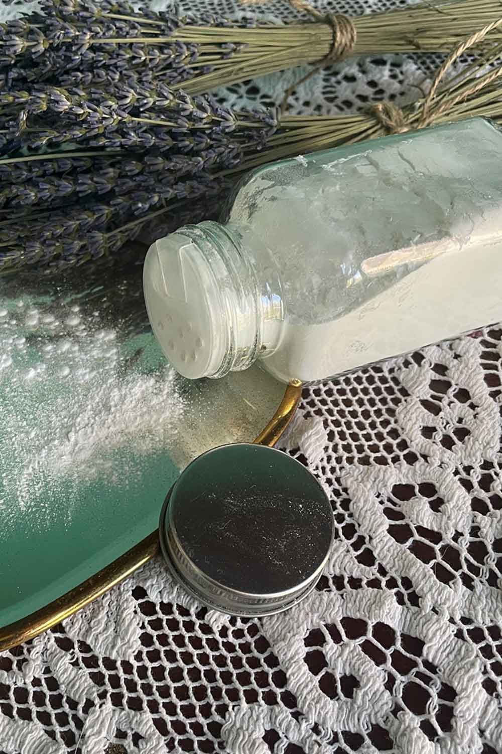 Vintage glass shaker of botanical dusting powder tipped onto a lace-covered vanity mirror with dried lavender sprigs — part of the Stillroom Dusting Powder series.