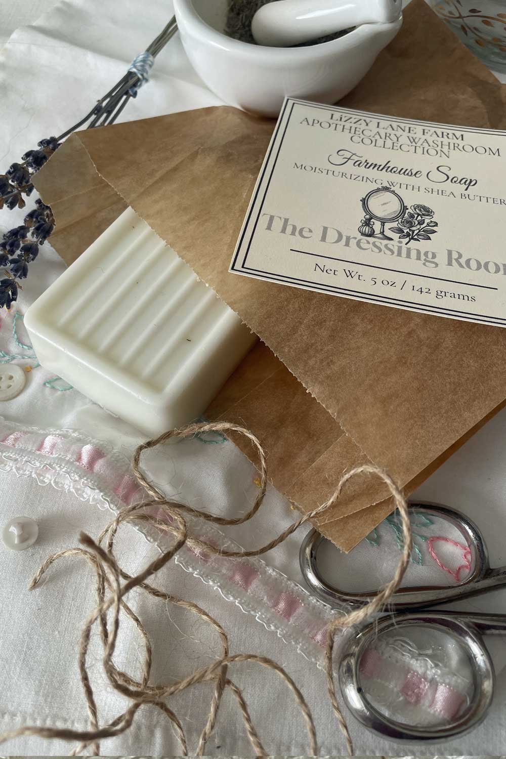 A creamy white bar of soap peeks out from kraft paper wrapping, tied with natural twine. The Dressing Room label rests atop, framed by delicate vintage lace and soft-hued buttons. Warm light spills across the scene, evoking a quiet, nostalgic moment.