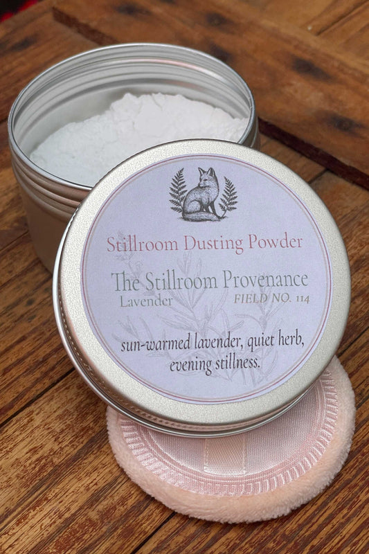 A tin of Stillroom Dusting Powder labeled 'The Stillroom Provenance' and 'Lavender,' shown open with white powder inside on a wooden surface, alongside a pale pink textured applicator puff.