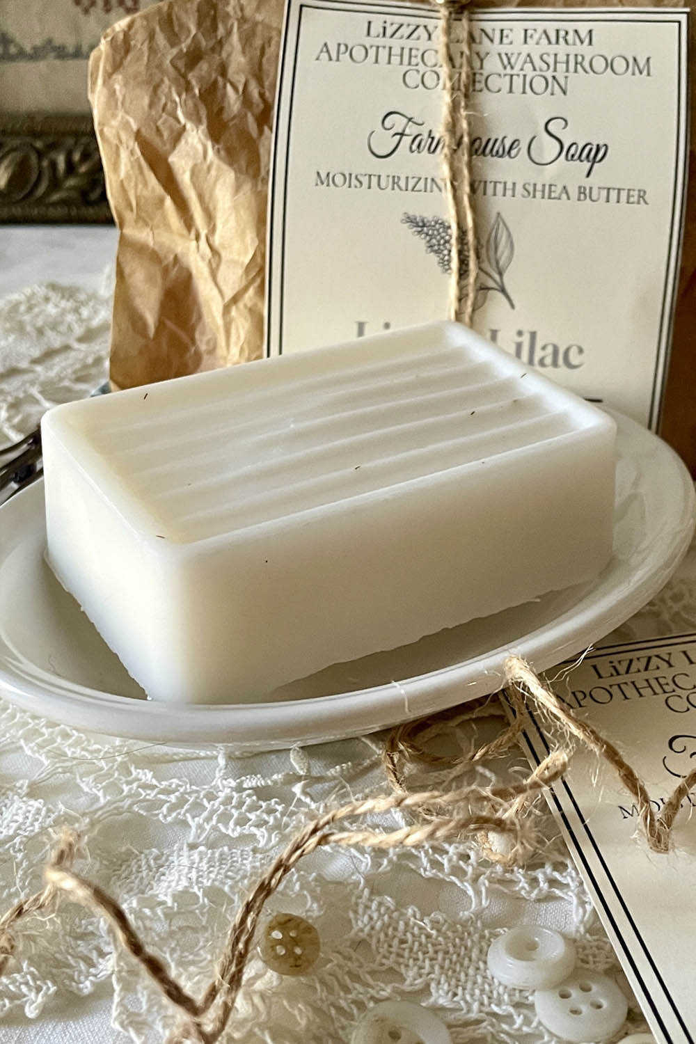 A creamy white bar of soap rests in a vintage porcelain soap dish, its grooved top catching the light. Beside it, a twirl of natural twine and a few vintage buttons add farmhouse charm. A peek of the label rests nearby, while a fully wrapped bar in kraft paper with twine and brass fastener stands in the background, showcasing the full packaging. The scene is softly lit, evoking a warm, nostalgic stillroom moment.