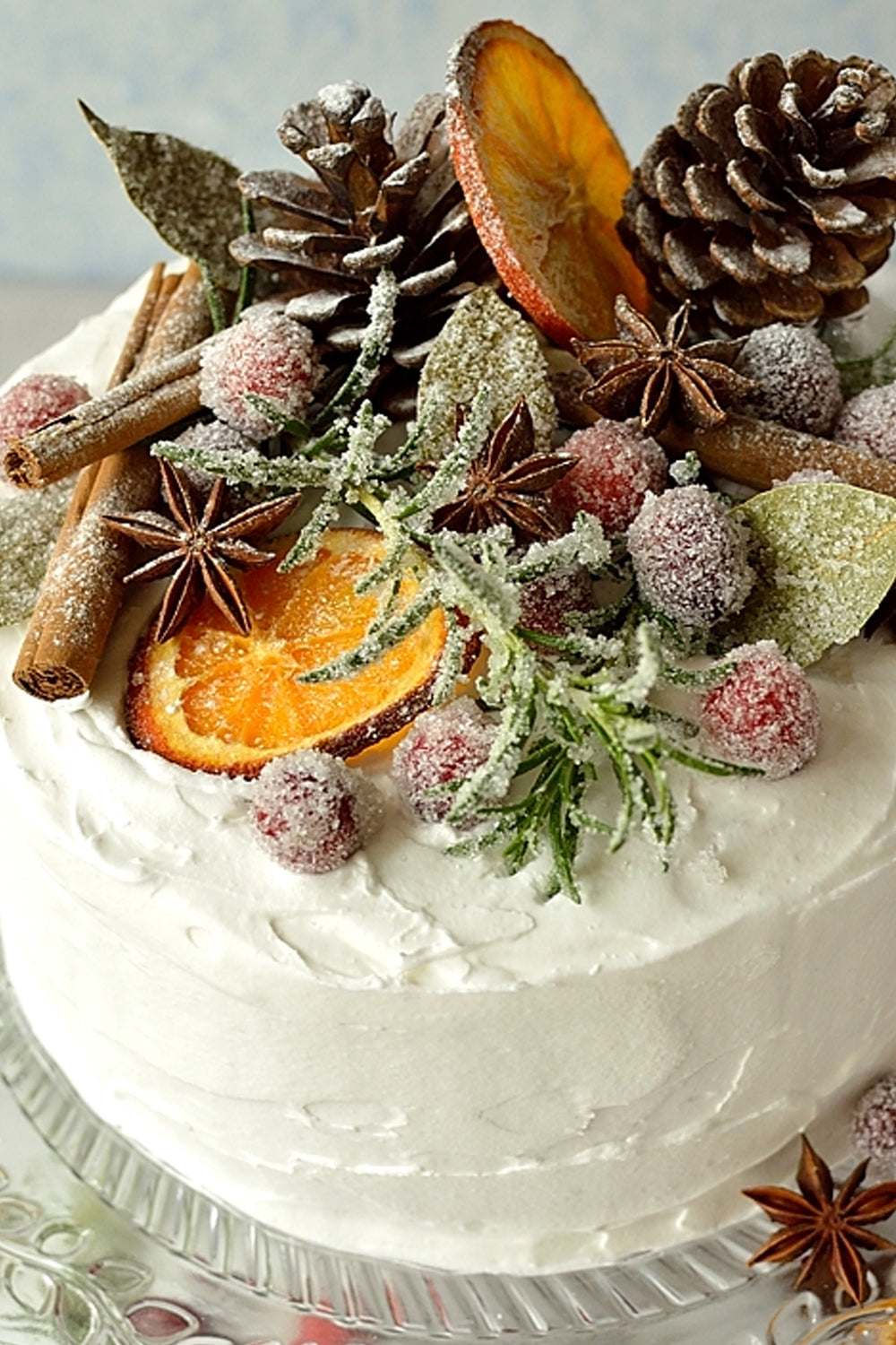 Frosted spice cake topped with sugared bay leaves, dried orange slices, star anise, cinnamon sticks, red berries, and a pine cone—rustic winter dessert styled with natural herbs and spices.