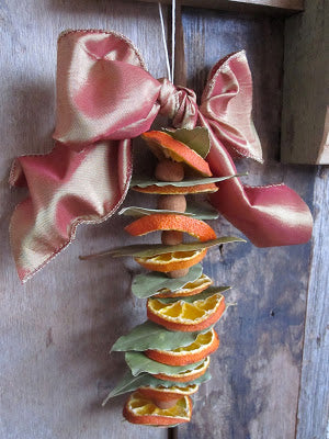Handmade fall decoration with strung dried orange slices, cinnamon sticks, and bay leaves, tied with a soft satin pink bow—natural autumn garland for home or kitchen décor.