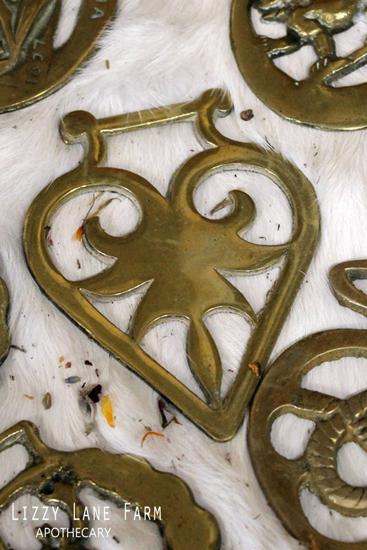 Assorted vintage horse harness brasses shown on a wood and lace backdrop with soft light. Designs vary; each brass shows gentle age and patina from years of use.