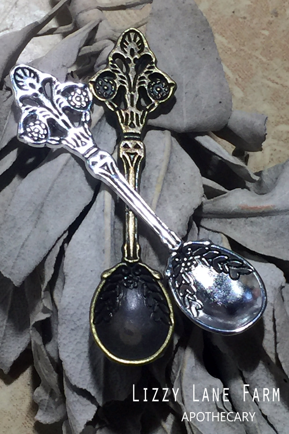 A close-up of two metal herbal spoons, one in silver tone and one in brass, elegantly resting on a bed of herbs. These petite spoons are part of our incense accessory kit, ideal for measuring and sprinkling loose incense or herbal blends.
