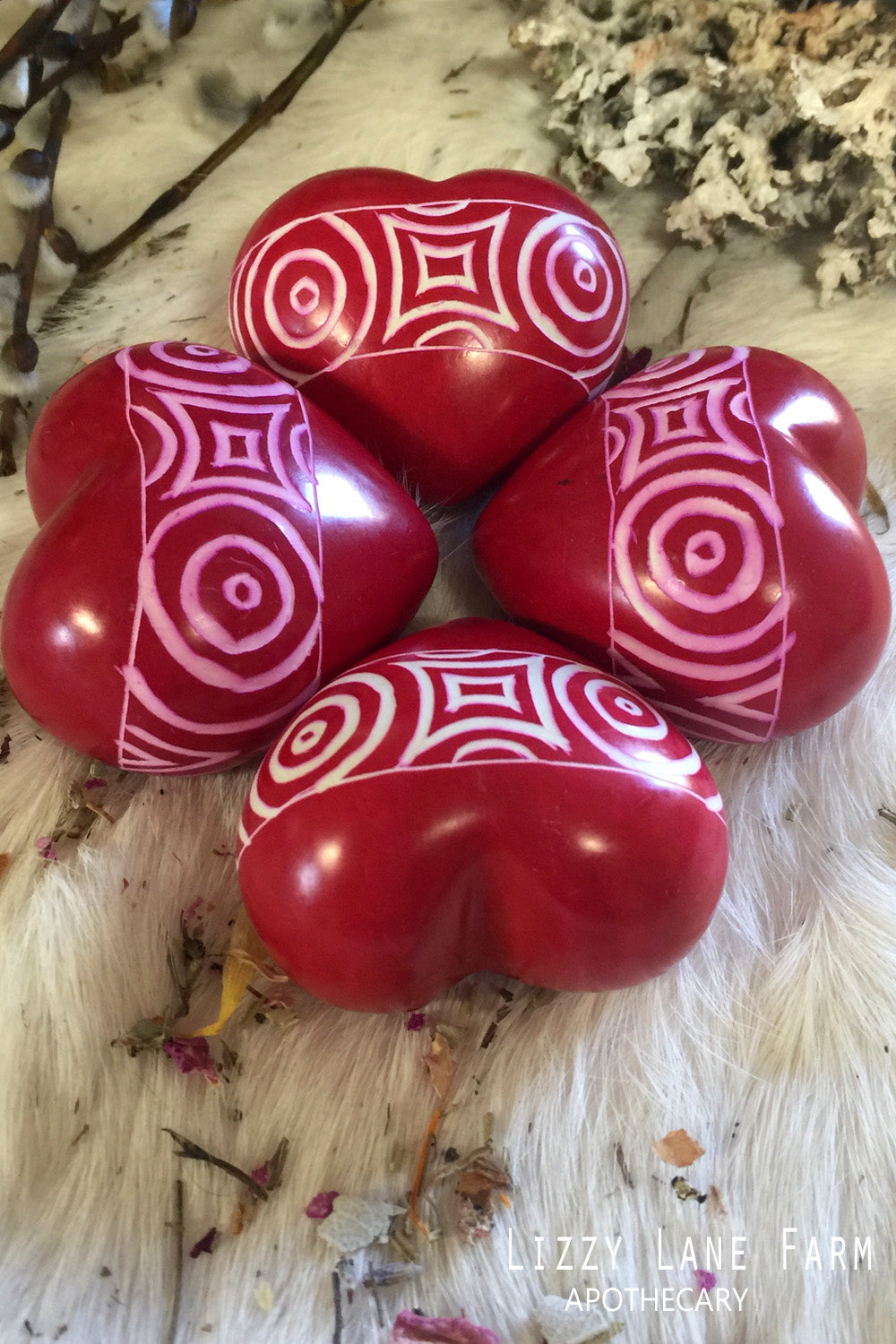 Bold red soapstone hearts with etched circular patterns, arranged in a star-like shape on soft fur. Surrounded by pussy willow and dried herbs in soft light.