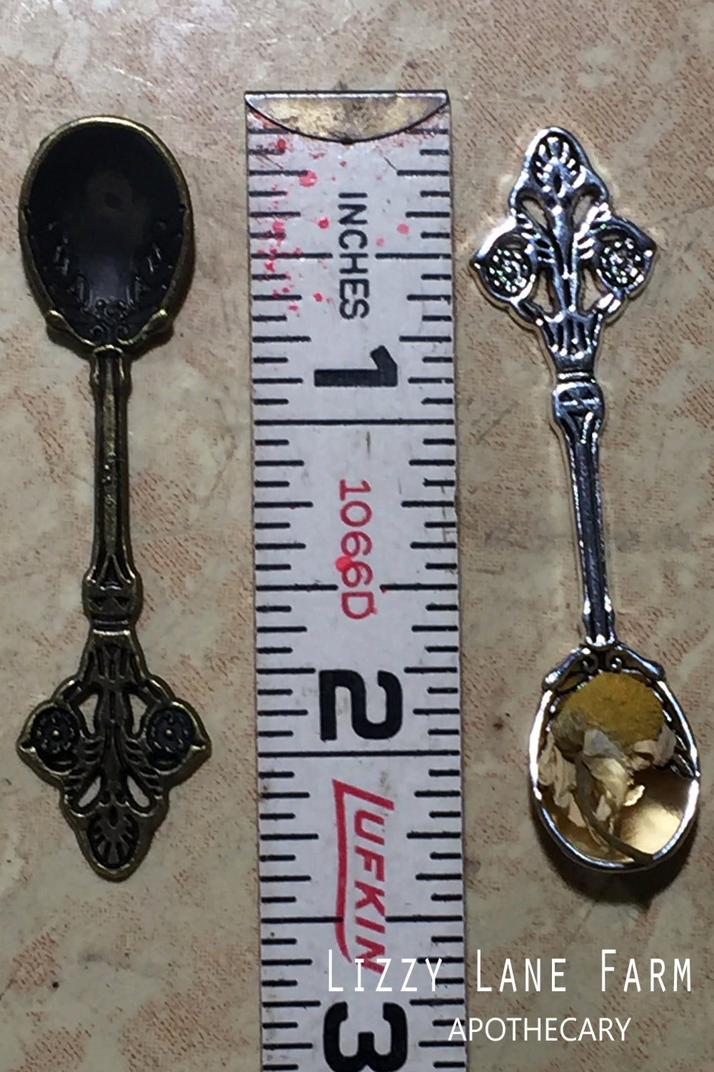 A close-up of two metal herbal spoons, one in silver tone and one in brass, elegantly resting on a bed of herbs. These petite spoons are part of our incense accessory kit, ideal for measuring and sprinkling loose incense or herbal blends.
