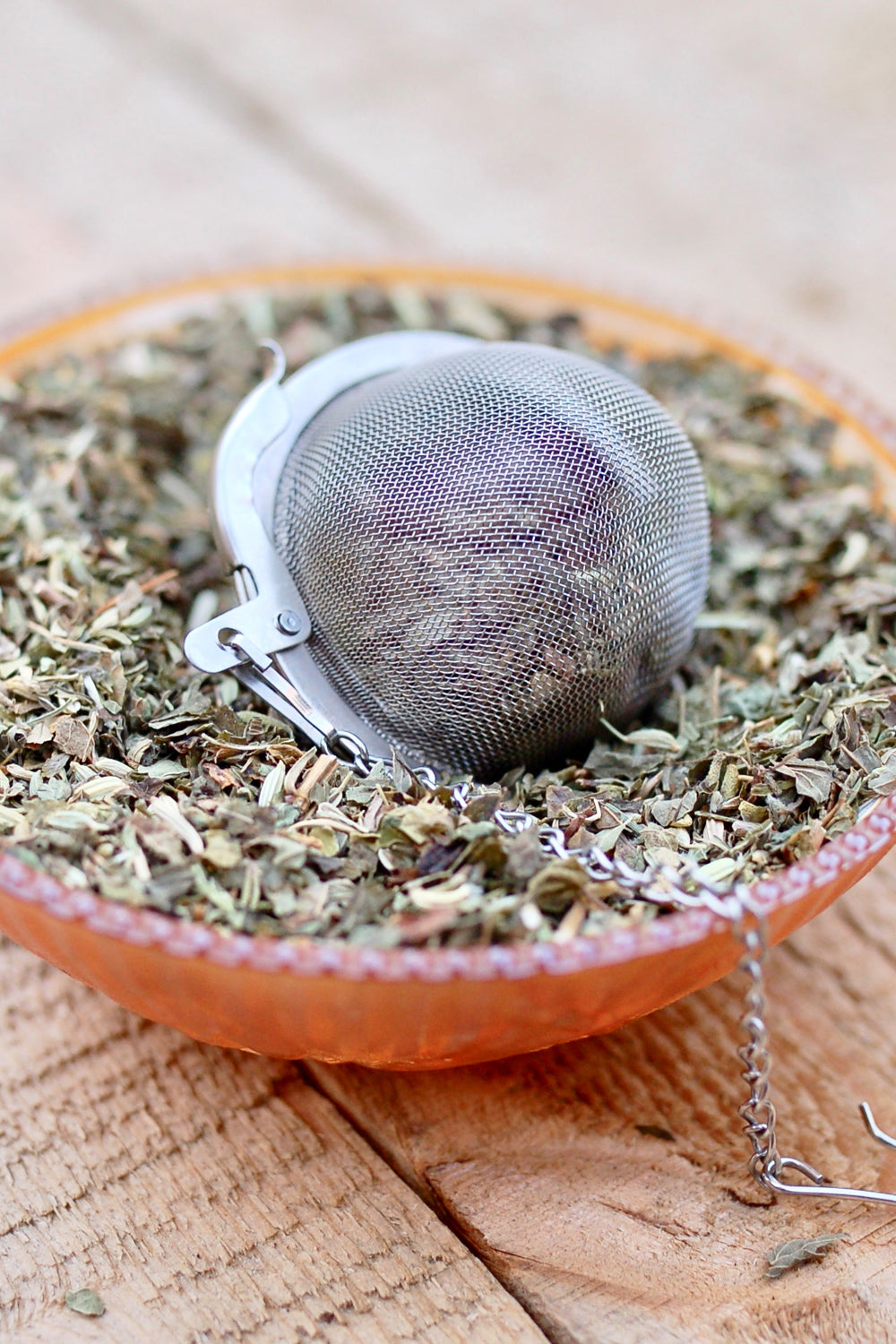 Stainless steel mesh tea ball resting in a bowl of loose leaf herbal tea on a rustic wood surface.