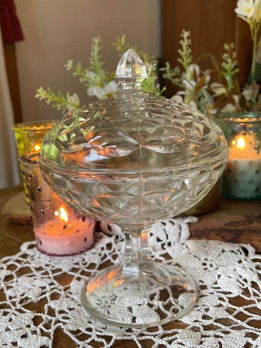 
Vintage pressed glass pedestal powder dish from Jeanette Glass Co., photographed on red and white crochet doily with lit candles in background – top view highlighting lid detail.