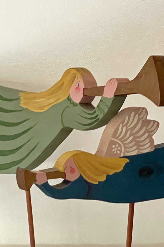 Close-up of two hand-painted wooden angels by Susan MacDonald, both with flowing blonde hair and rosy cheeks, holding trumpets. The larger angel wears a green dress with wing detail, while the smaller angel below wears a dark teal dress with painted white wings.