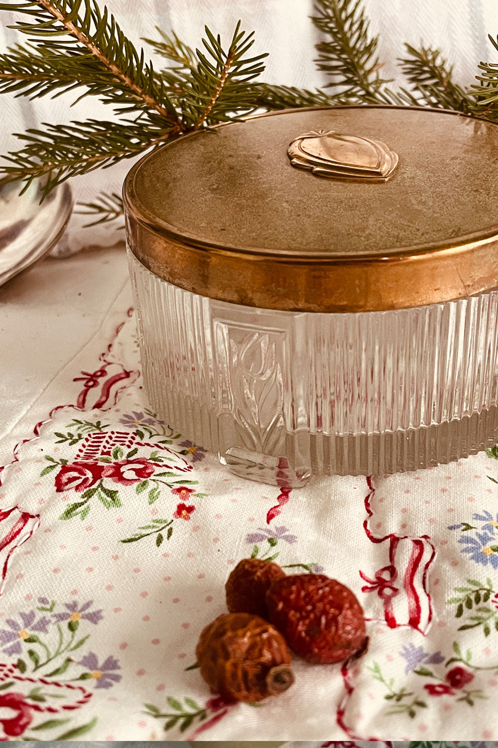 Vintage pressed-glass powder dish with raised floral design and brass Art Deco lid, photographed on a floral quilt with evergreen sprigs in soft natural light.
