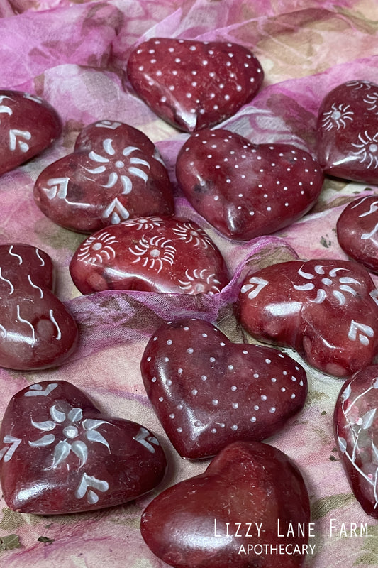 Small red hand-carved soapstone heart resting on linen beside dried flowers and a folded note. Warm tone and soft texture visible under natural light.