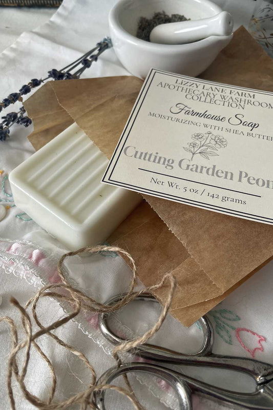 A creamy white farmhouse soap bar of cutting garden is partially unwrapped in kraft paper, its grooved top just visible. A soft twirl of twine rests nearby, with a simple black-and-white label lying on top. Vintage lace and buttons frame the scene, evoking the warmth of a country kitchen.