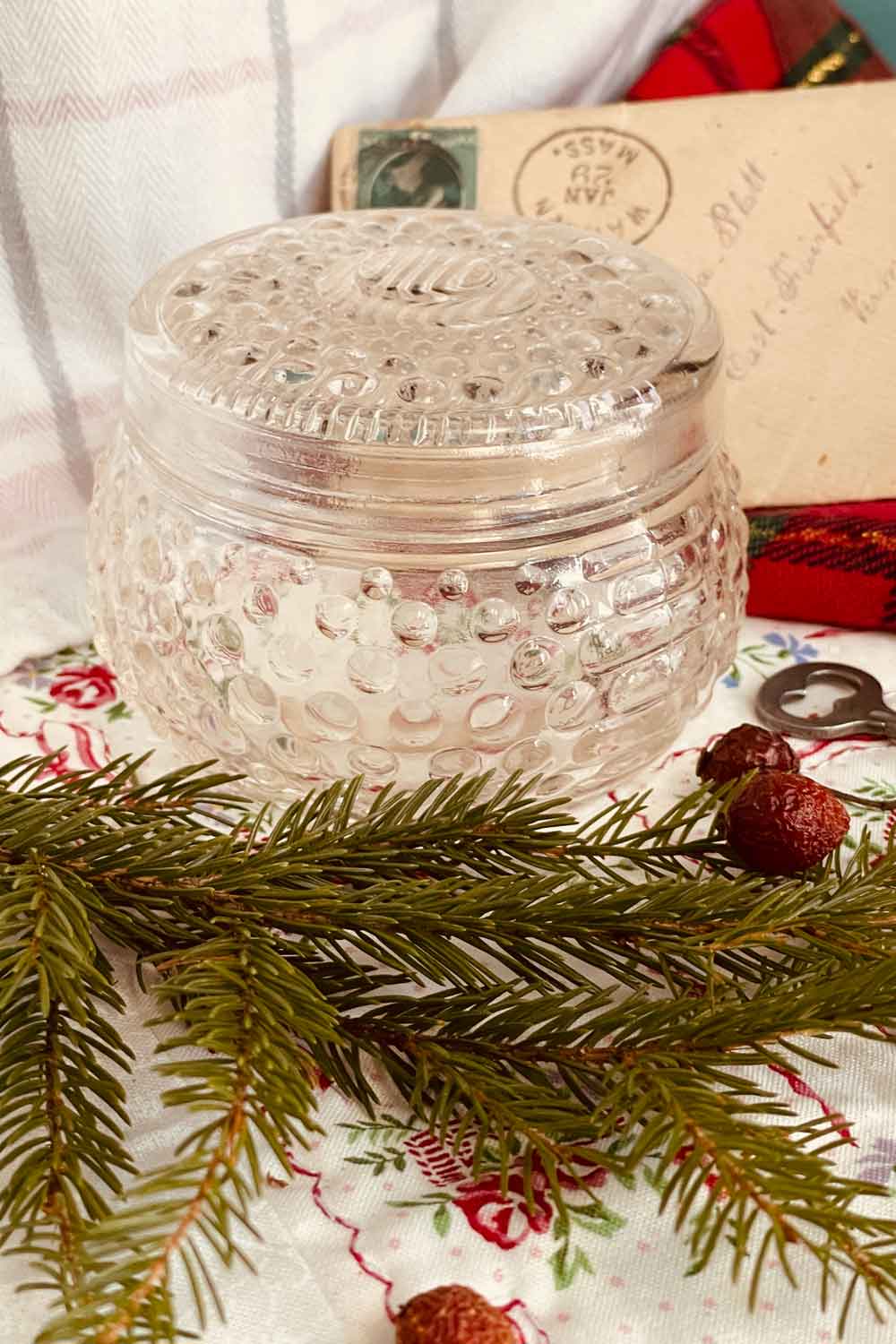 Open hobnail powder dish with matching glass lid beside it, photographed on a vintage floral cloth with winter greenery.