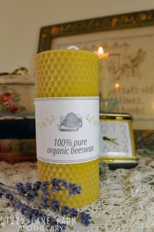 A hand-poured organic beeswax pillar candle with a molded honeycomb design sits on a vintage wooden dresser, exuding rustic charm. In the softly blurred background, a second beeswax candle flickers warmly, casting a golden glow. The natural honey scent of pure beeswax fills the air, creating a cozy and inviting atmosphere. This handmade beeswax candle, crafted from locally sourced New Hampshire beeswax, offers a clean, steady burn and timeless farmhouse elegance.
