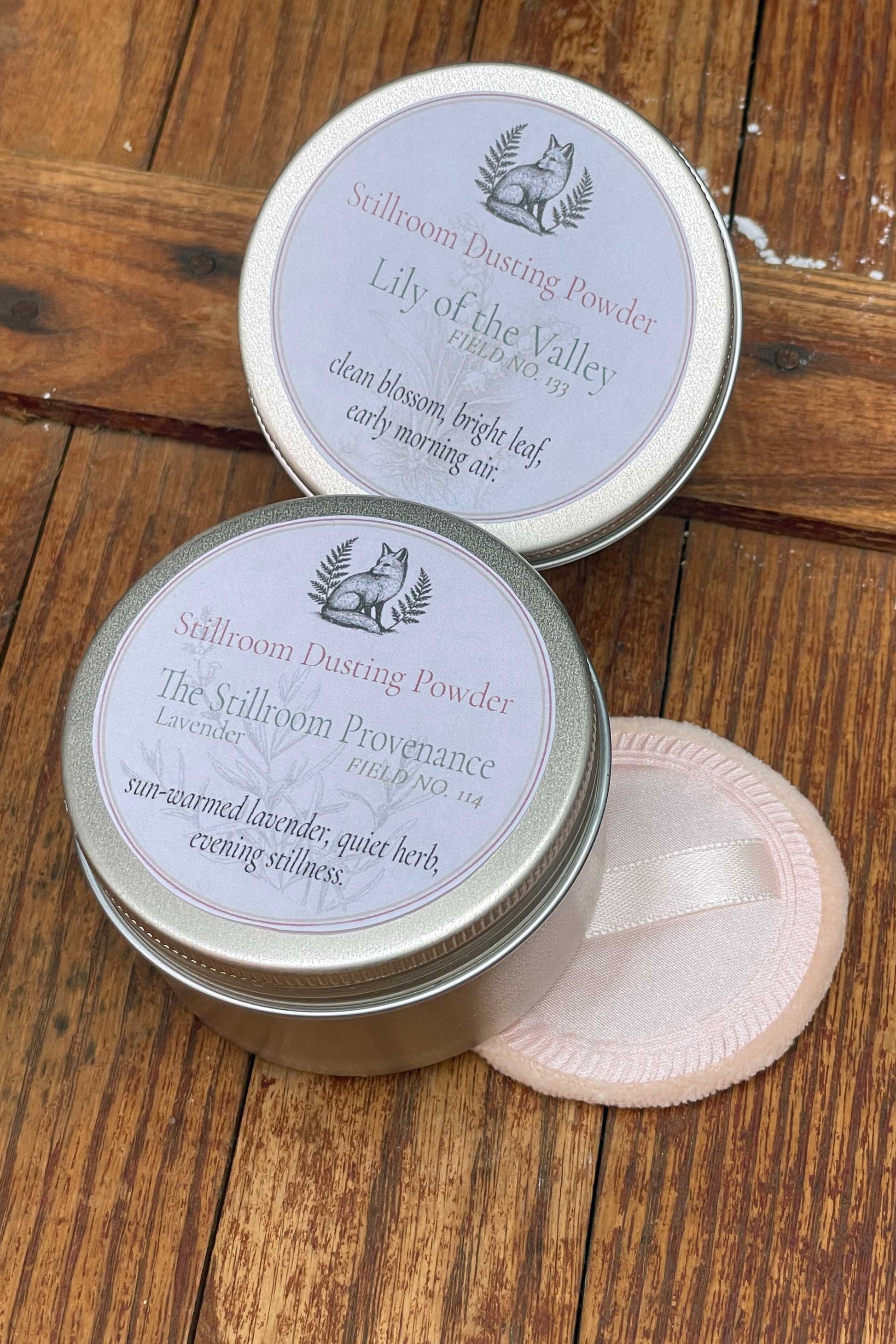 Two round metal tins of sillaroom dusting powder with pale lavender labels, placed on a rustic wooden surface, with a light pink round puff packet beside them.