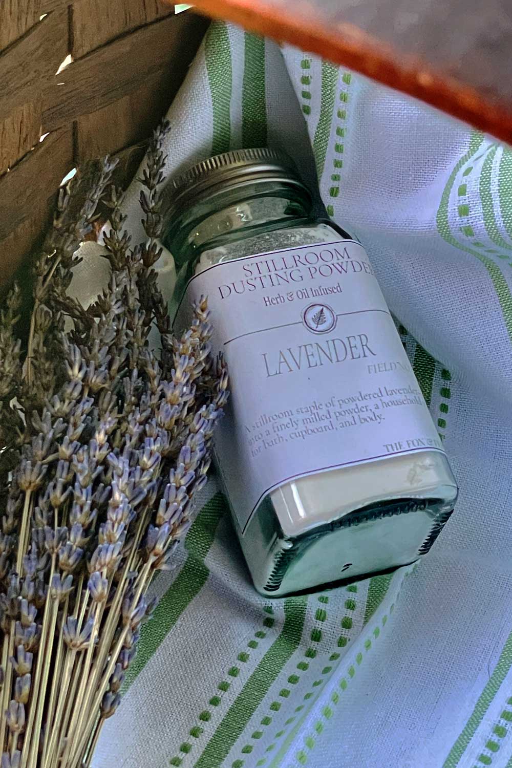 A glass jar of Stillroom Dusting Powder, Lavender, resting on a green and white striped kitchen towel next to dried lavender sprigs.