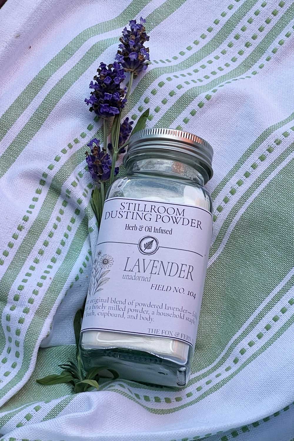 An apothecary glass powder shaker labeled 'Stillroom Dusting Powder' with lavender flowers placed on top, set on a white and green striped fabric background.