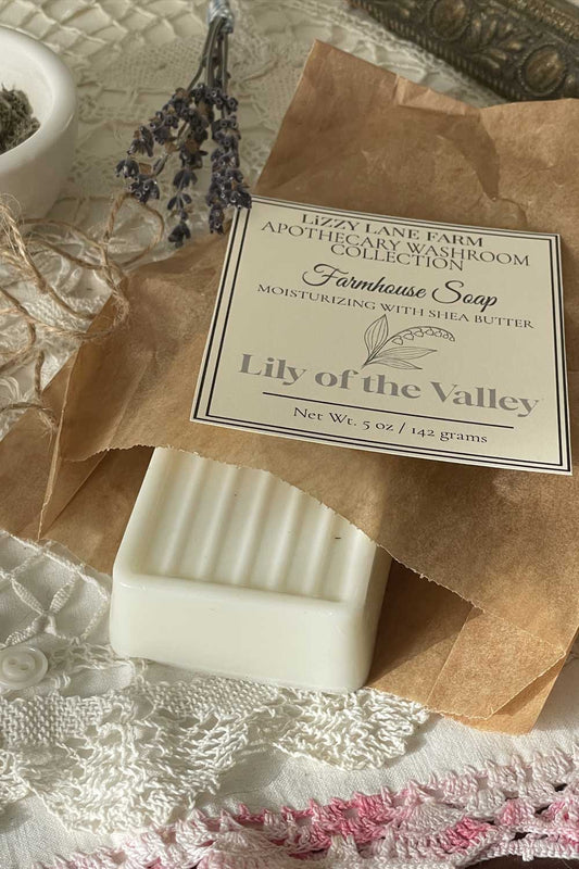 A creamy white bar of soap wrapped in kraft paper, partially unwrapped to reveal soft grooves and smooth texture. The label rests on top of the folded wrapping, with twine loosely curled nearby. Delicate vintage lace and ivory buttons frame the scene, evoking the quiet charm of a farmhouse vanity. Sunlight warms the entire image with a gentle glow.