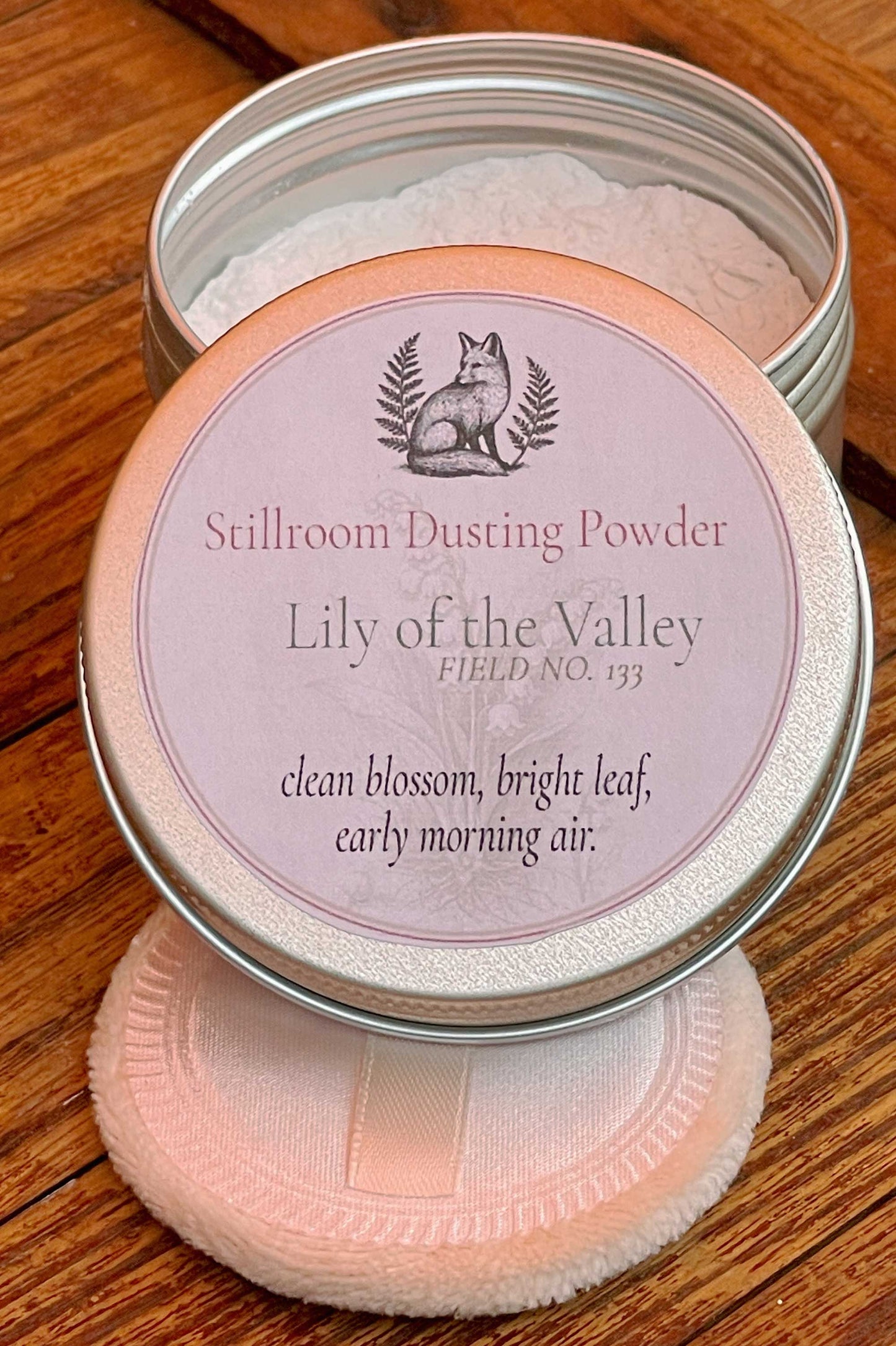 A round metal tin of Stillroom Dusting Powder from Lily of the Valley, shown open with the lid resting on top and a soft pink dusting pad placed in front, all on a wooden surface.