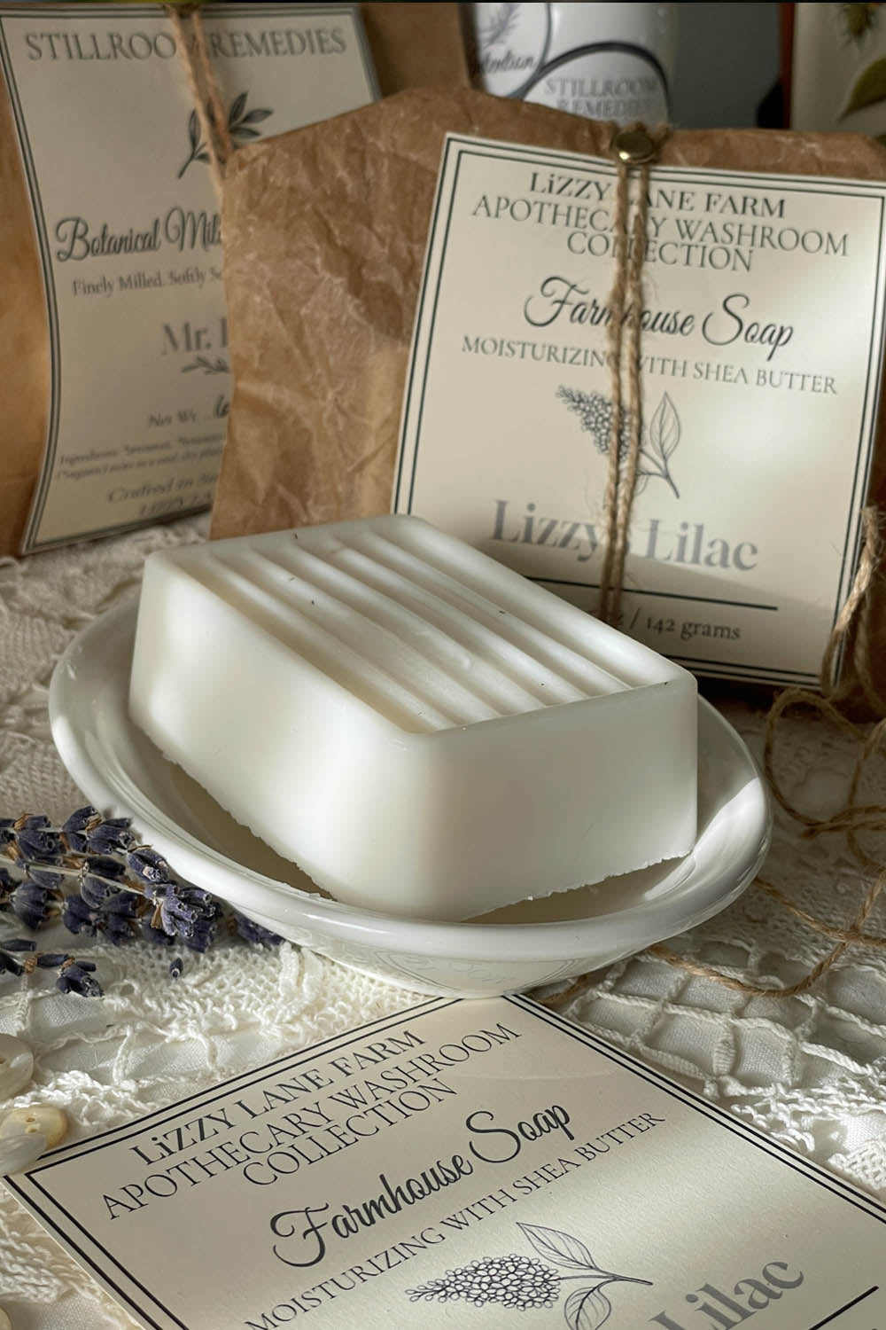 A handcrafted white soap bar with elegant grooves sits in a vintage white dish, partially unwrapped to reveal its texture. Behind it, kraft paper packaging is tied with twine and sealed with a brass fastener. The label reads “Lizzy’s Lilac – Apothecary Washroom Collection” from Lizzy Lane Farm. Dried lavender sprigs, ivory buttons, and vintage lace add a nostalgic farmhouse touch.