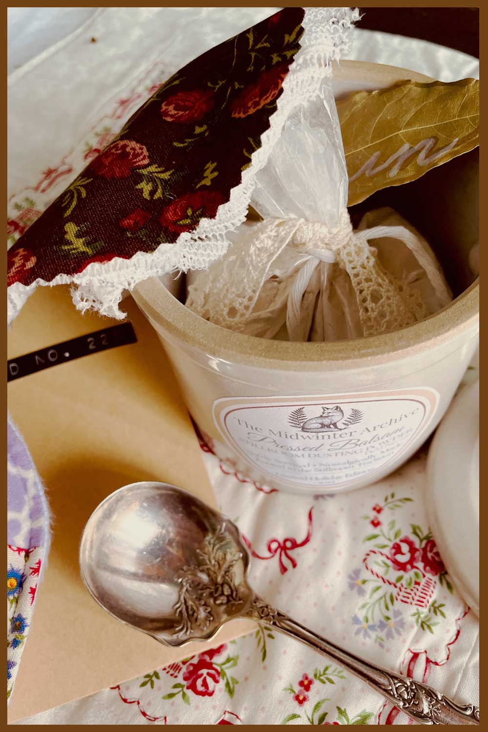 Inside the Midwinter Powder Crock — Pressed Balsam dusting powder wrapped in lace and cheesecloth, with a gold-edged bay leaf marked “turn.”