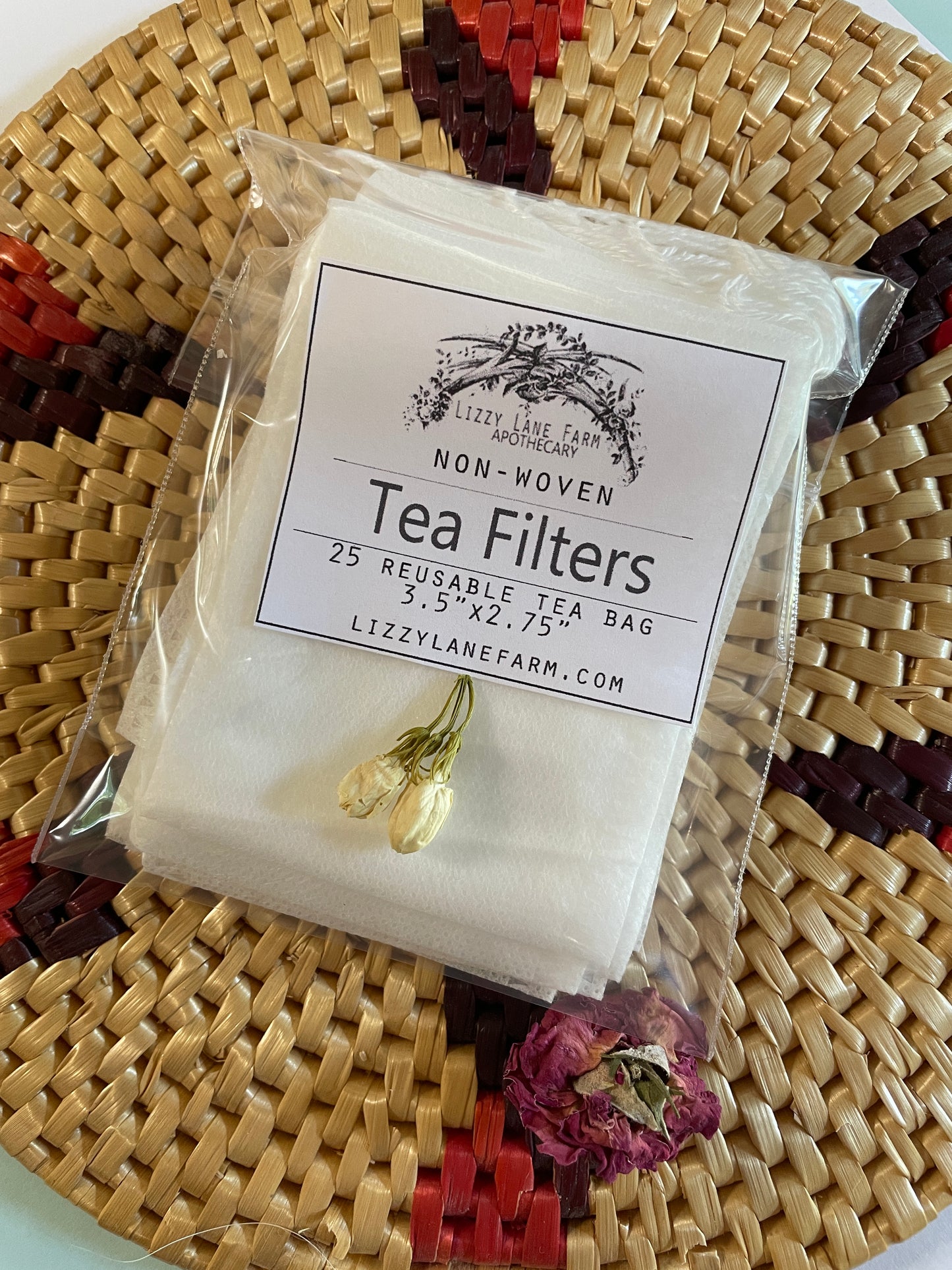 A package of reusable non-woven tea bags resting on a woven natural fiber mat. The tea bags are visible through the packaging, showcasing their soft texture and drawstring closure. The earthy tones of the mat complement the eco-friendly, farmhouse-inspired aesthetic.