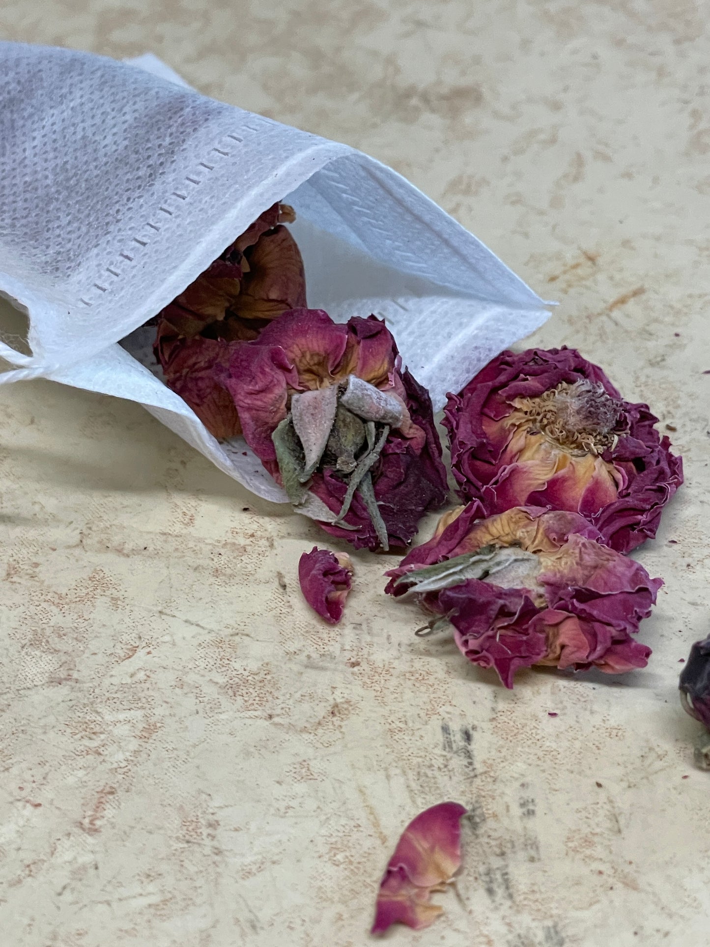 An open reusable non-woven tea bag with drawstrings spread apart, spilling dried rose buds onto a woven natural fiber mat. The delicate petals contrast with the soft texture of the bag, highlighting its eco-friendly and functional design.