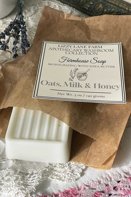 A creamy, white handmade bar of soap rests partially unwrapped in soft kraft paper, revealing its smooth texture and traditional grooved top. Twine curls nearby, and a printed label lies neatly across the wrapping. The background features vintage lace and a few scattered buttons, creating a warm, nostalgic farmhouse scene.